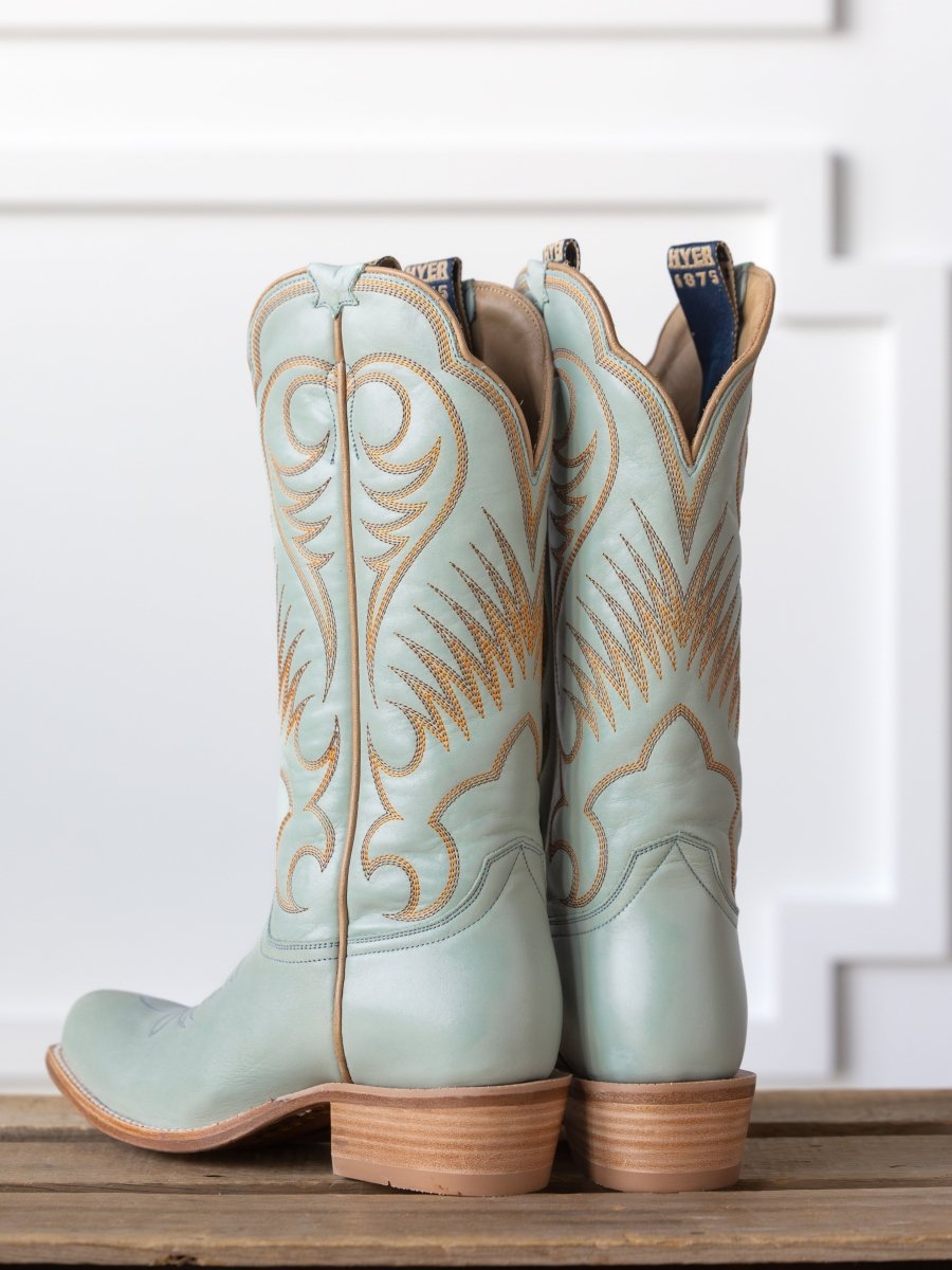 A pair of The Hyer Leawood Boot {Robin's Egg} from Savannah Sevens western life{&}style with tan stitching sit side by side on a wooden surface, set against a white paneled wall.
