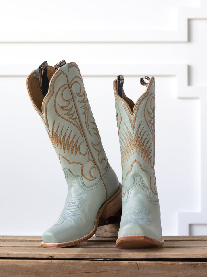 A pair of The  Hyer Leawood Boot {Robin's Egg} by Savannah Sevens western life{&}style, light blue with tan stitching and soles in classic Western leather style, stands on wood against a white patterned wall.