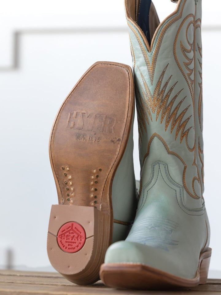 A close-up of two Savannah Sevens western life{&}style  Hyer Leawood Boots in Robin's Egg—with tan stitching and a branded leather sole—showcases Western craftsmanship and Goodyear welt construction, both resting on a wooden surface.