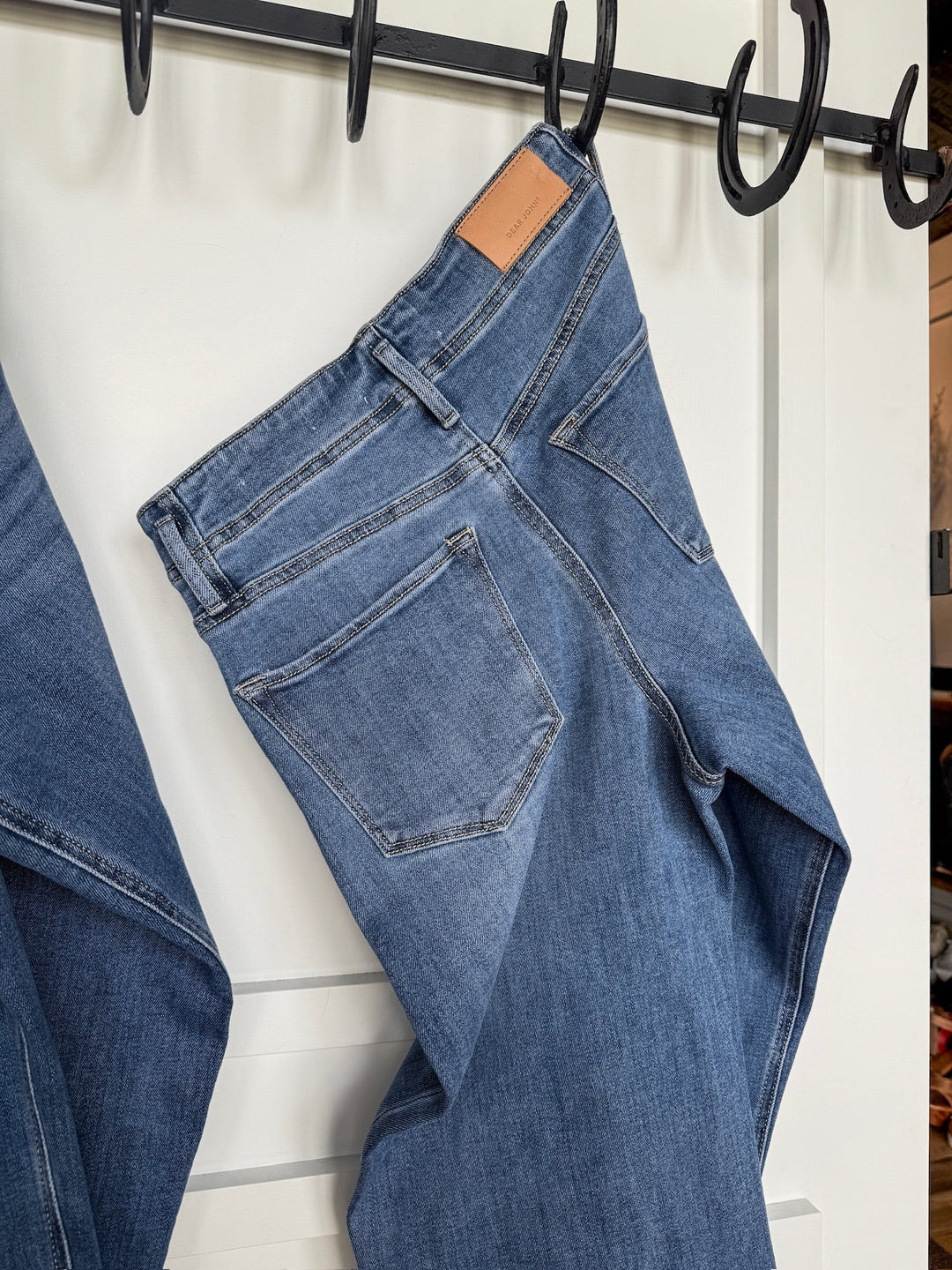 A pair of Savannah Sevens western life{&}style The Leddy High Rise Wide Leg Jeans with a brown leather patch on the waistband hangs over a black metal hook on a white wall.