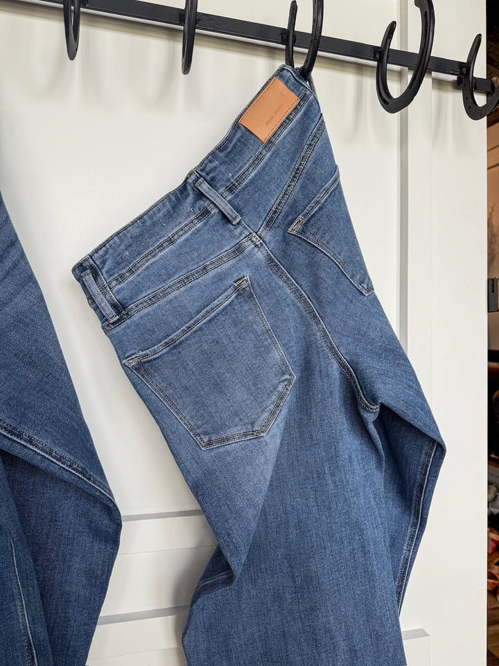 A pair of Savannah Sevens western life{&}style The Leddy High Rise Wide Leg Jeans with a brown leather patch on the waistband hangs over a black metal hook on a white wall.
