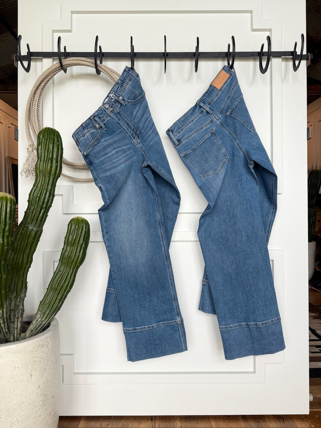 Two pairs of The Leddy High Rise Wide Leg Jeans by Savannah Sevens western life{&}style hang on black hooks against a white paneled wall, with a coiled rope and tall potted cactus showcasing their ultra high rise wide leg stretch denim design.