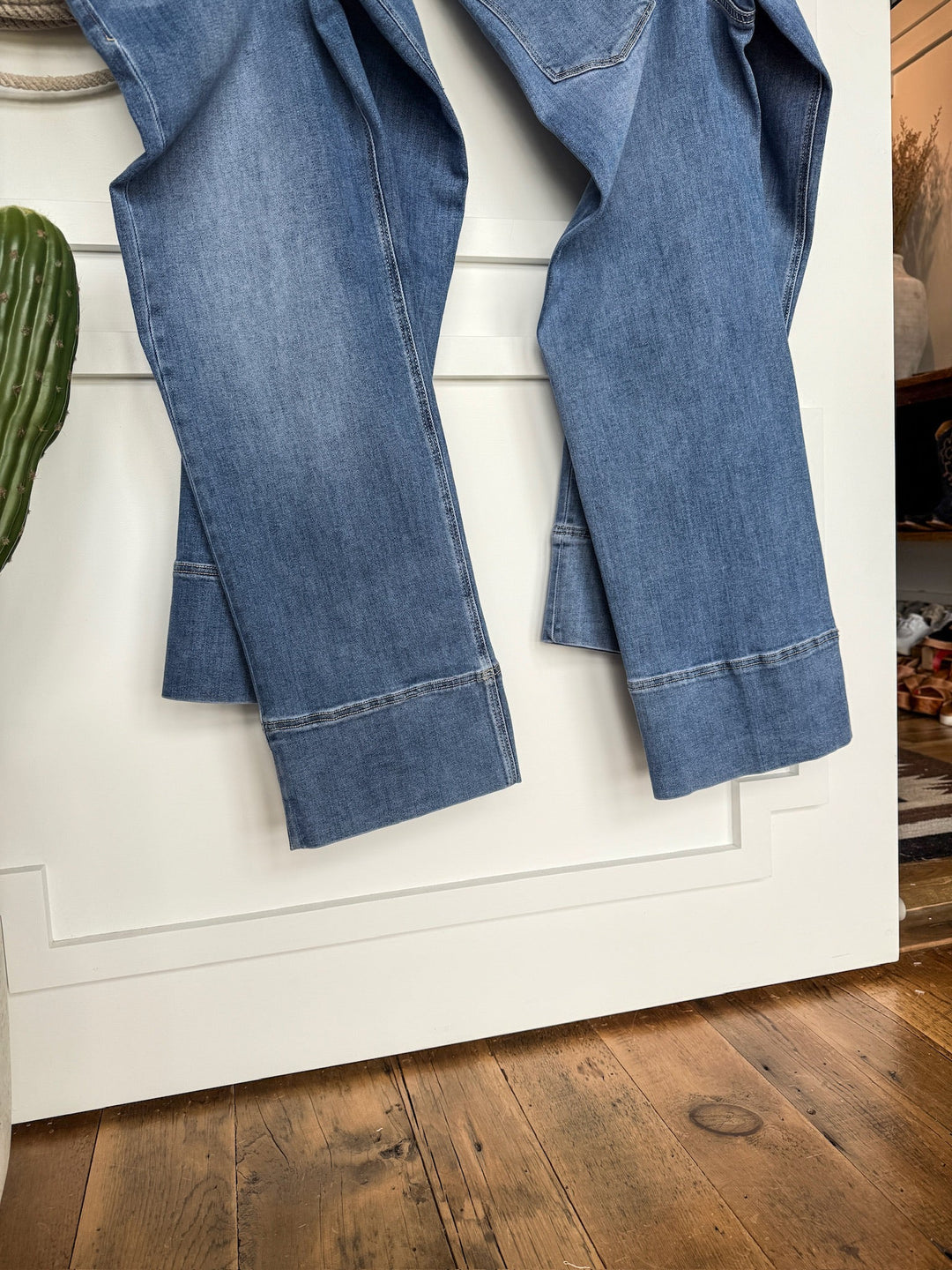 A pair of The Leddy High Rise Wide Leg Jeans by Savannah Sevens western life{&}style hangs over a white door beside a green cactus on wood flooring, adding style to the cozy, sunlit room in the background.