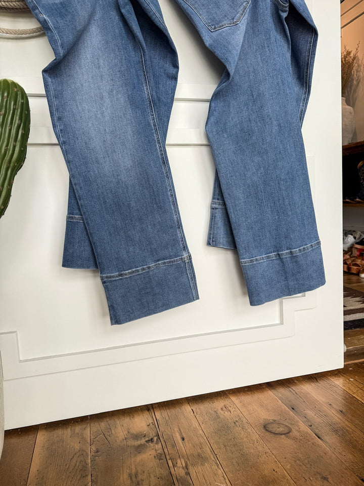 A pair of The Leddy High Rise Wide Leg Jeans by Savannah Sevens western life{&}style hangs over a white door beside a green cactus on wood flooring, adding style to the cozy, sunlit room in the background.