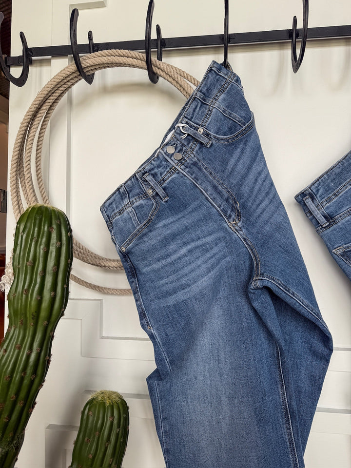 A pair of Savannah Sevens western life{&}style The Leddy High Rise Wide Leg Jeans hangs on black hooks against a white wall, with a green cactus and coiled rope nearby. These ultra high rise jeans are made from stretchy denim for comfort.