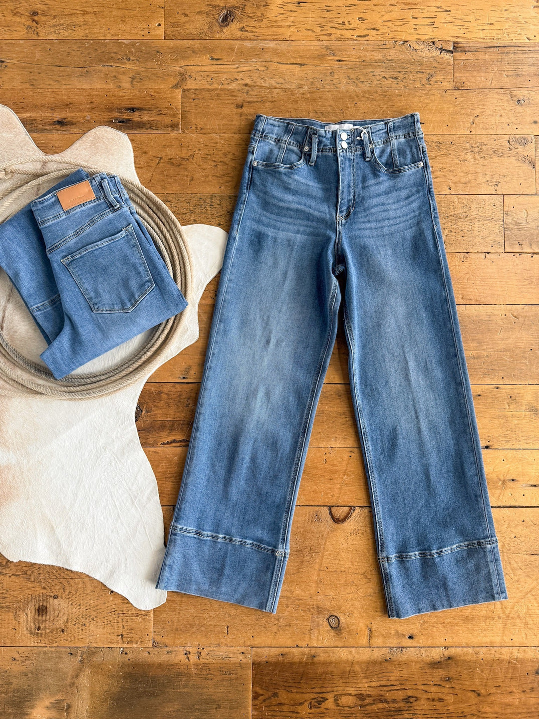 Two pairs of blue jeans are displayed on a wooden floor. One is folded on a white animal hide, while the other—The Leddy High Rise Wide Leg Jean by Savannah Sevens western life{&}style—lies flat, showing its stretchy denim and stitched cuffs.