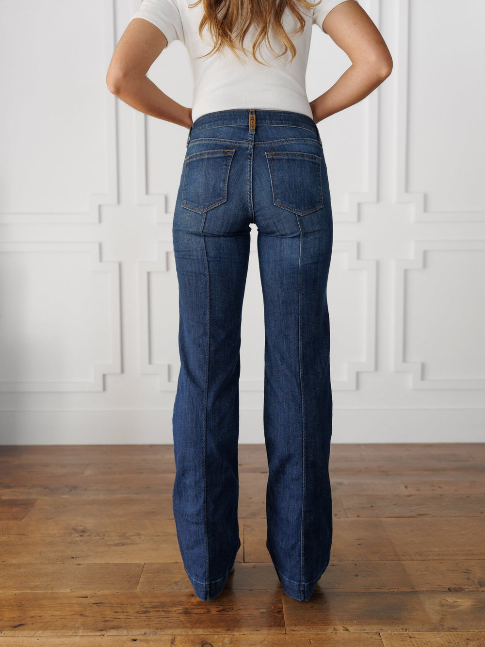 A woman with long hair, in a white short-sleeve top, stands by a white wall on a wooden floor wearing The Ledger Perfect Rise Trouser Jean *Exclusive* in medium dark wash, featuring back pockets and center seams.