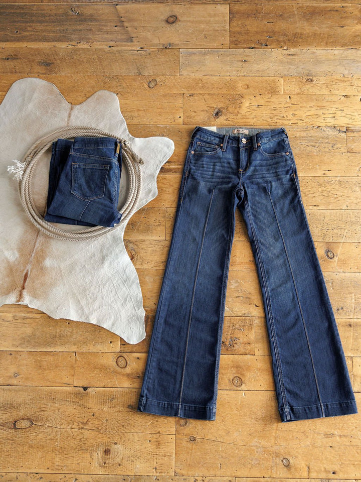 The Ledger Perfect Rise Trouser Jean *Exclusive* in dark blue is laid flat on a wooden floor, while a folded pair rests on a light animal hide with a coiled rope on top.