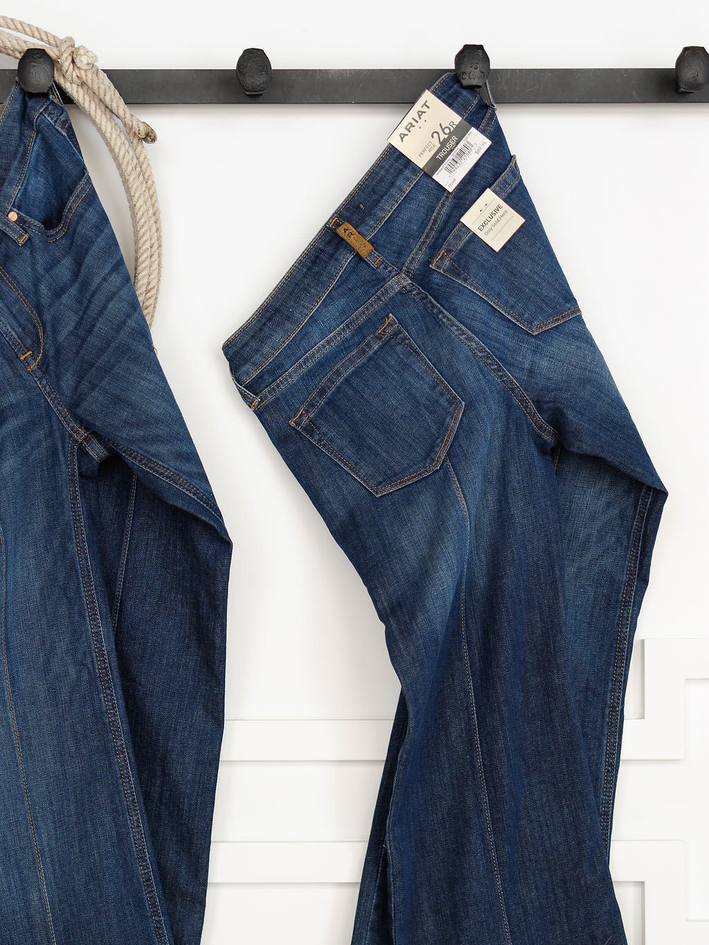 A pair of The Ledger Perfect Rise Trouser Jean *Exclusive* in a medium dark wash hangs on a black wall hook tags visible on the waistband, set against a white background.