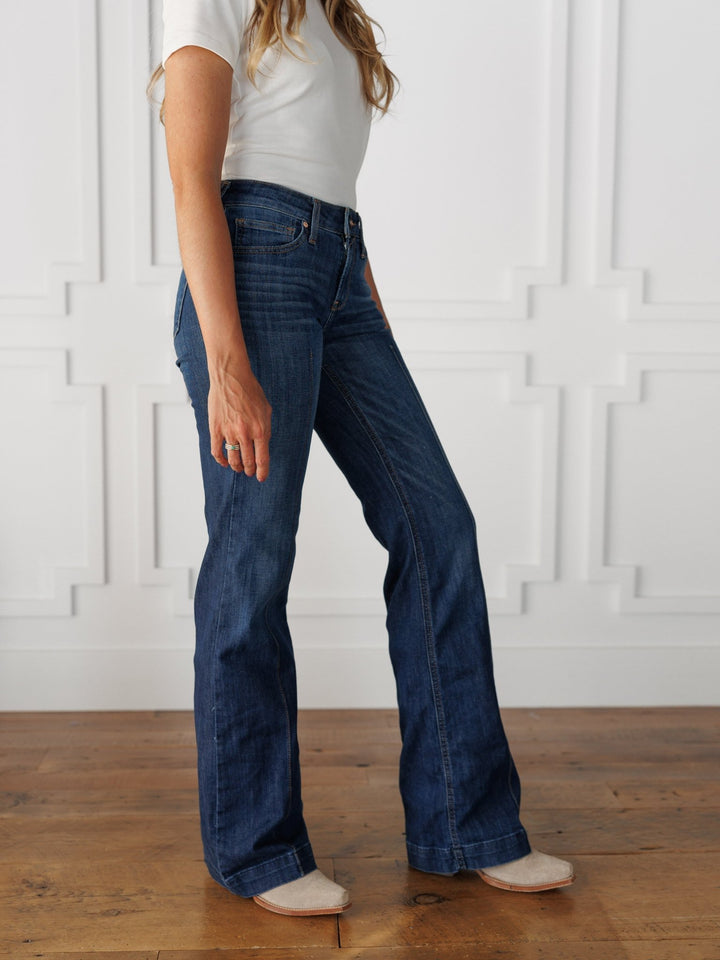 A woman stands indoors, posed sideways with her left arm relaxed, wearing a white short-sleeve top and The Ledger Perfect Rise Trouser Jean *Exclusive* in a medium dark wash, paired with light-colored shoes against a white paneled wall.