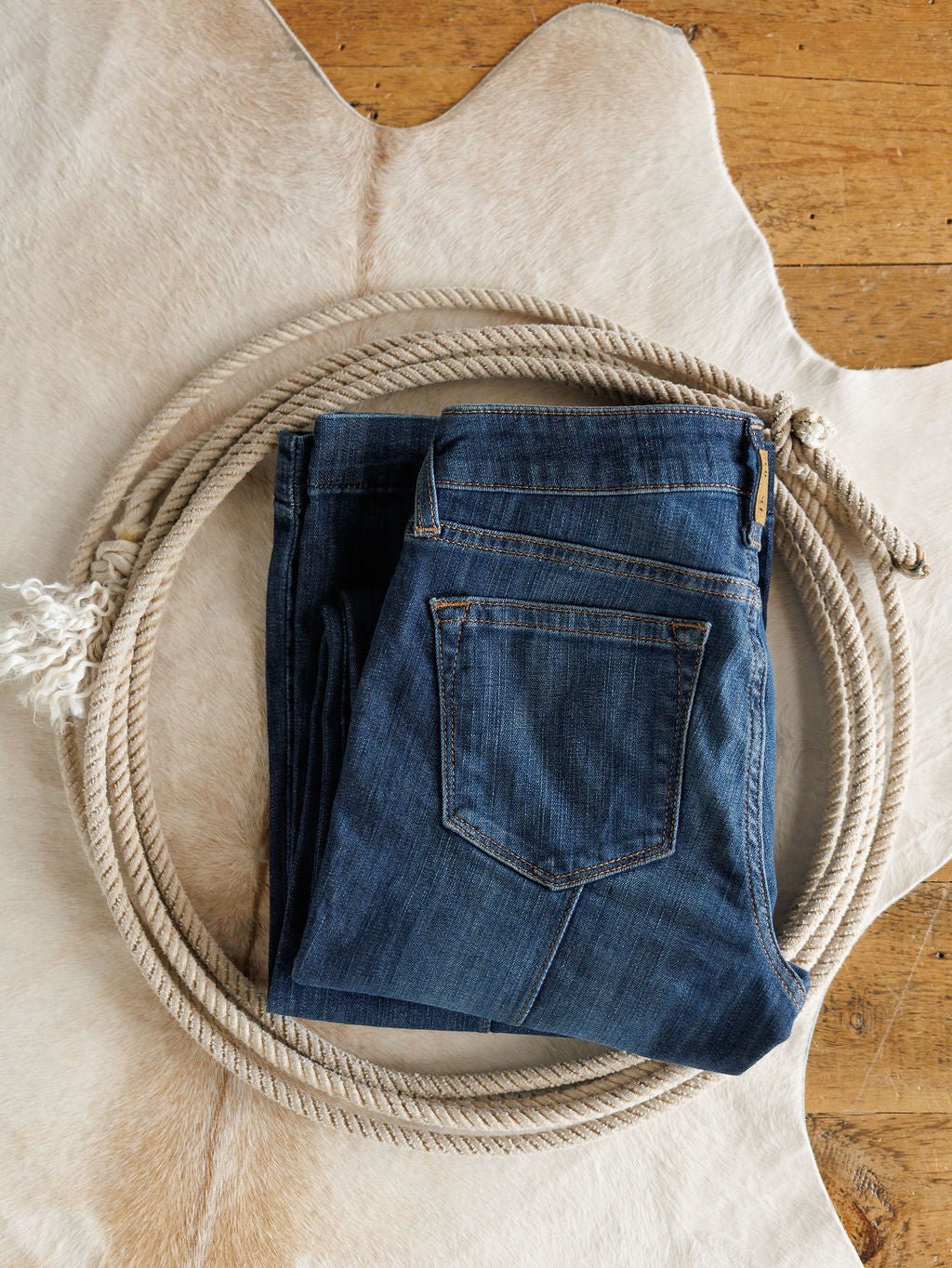 The Ledger Perfect Rise Trouser Jean *Exclusive* in a medium dark wash is folded inside a coiled rope on a light animal hide rug, with the wooden floor visible underneath.