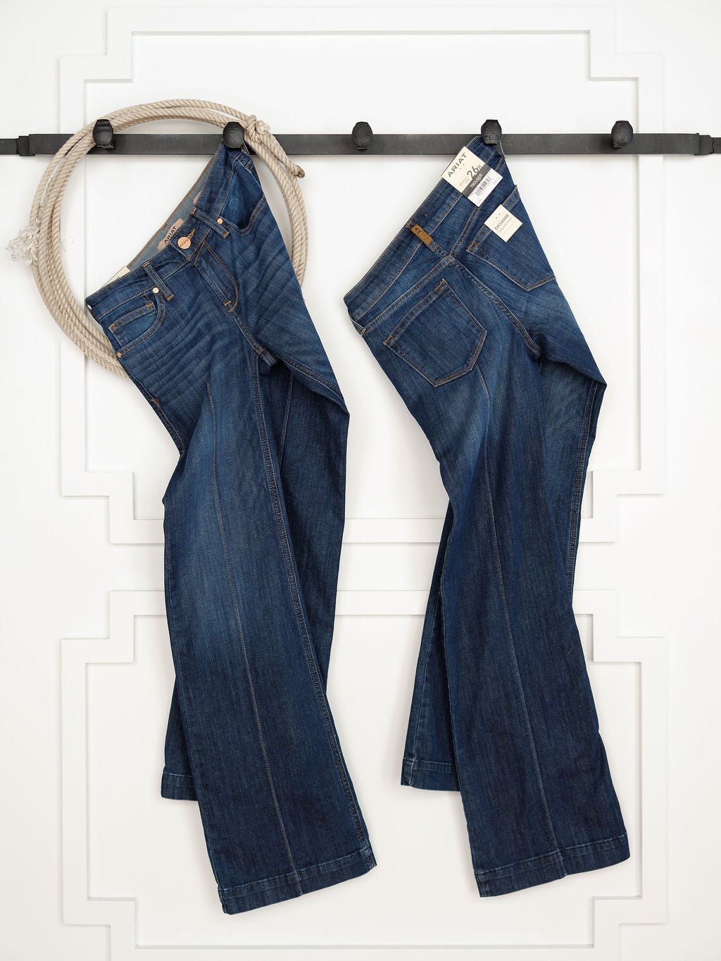 Two pairs of The Ledger Perfect Rise Trouser Jean *Exclusive* in a medium dark wash hang from a black rod on a white wall with decorative molding.