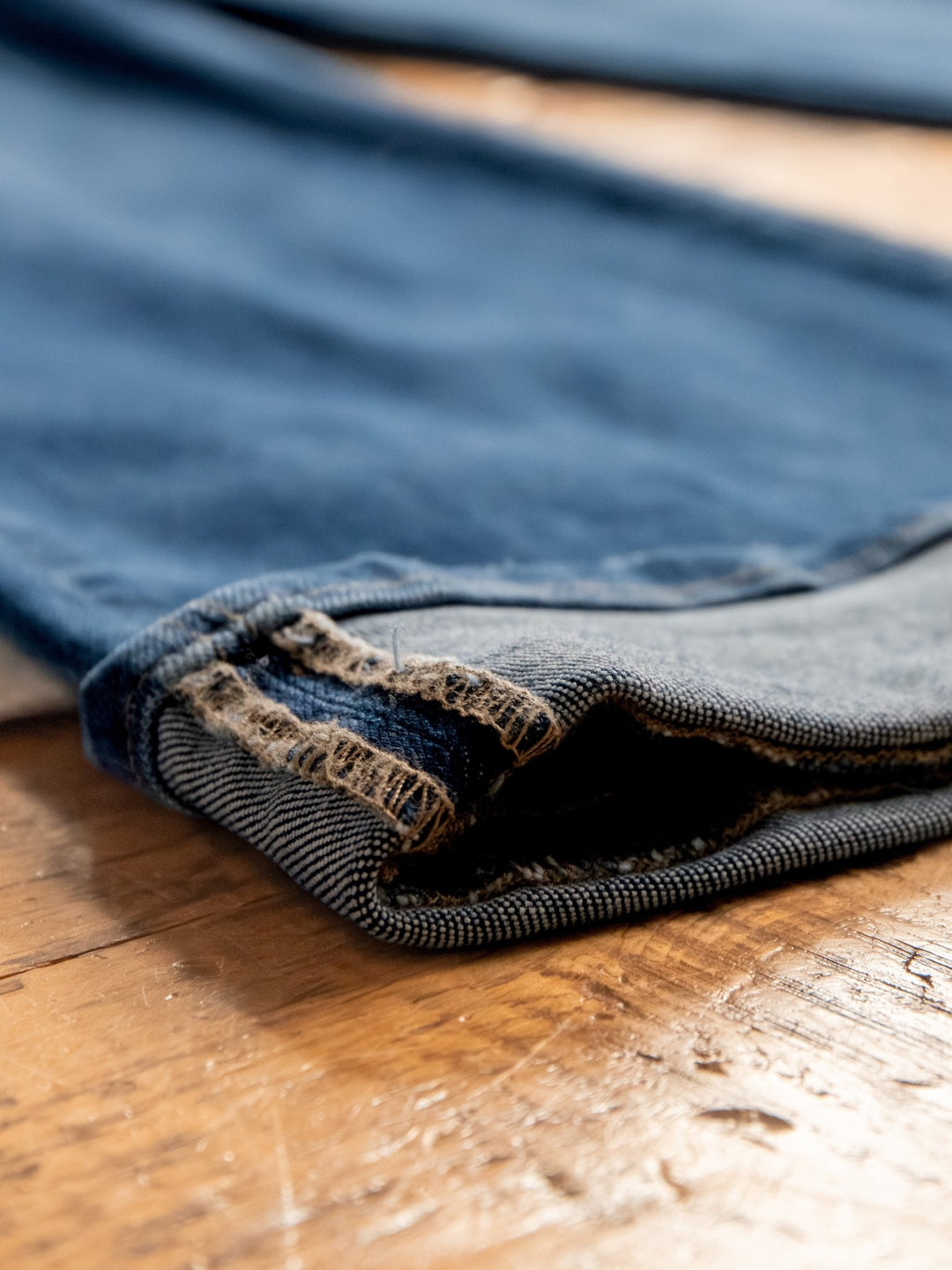 A close-up of the frayed, worn hems of The Lenni High Rise Slim Wide Leg Jean by Savannah Sevens western life{&}style in a medium dark wash, displayed on a wooden surface.