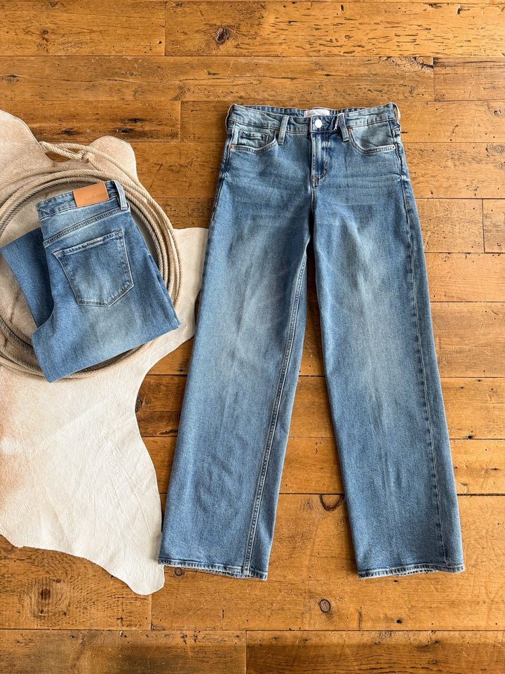 A pair of medium wash jeans lies flat on a wooden floor beside another folded pair of Savannah Sevens western life{&}style The Lennox Premier Rise Relaxed Straight Leg Jeans placed on a white cowhide rug and coiled rope.