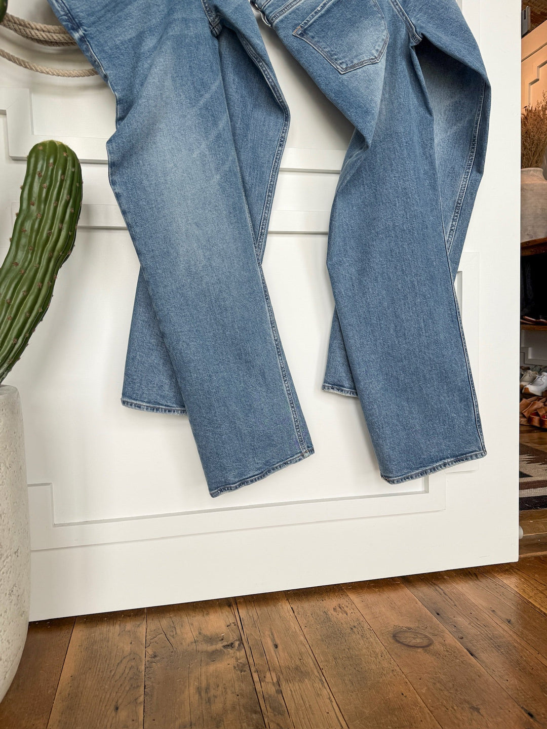 A pair of Savannah Sevens western life{&}style The Lennox Premier Rise Relaxed Straight Leg Jeans in medium wash hangs over a white door beside a potted cactus on a wooden floor.