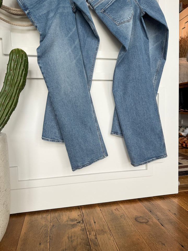 A pair of Savannah Sevens western life{&}style The Lennox Premier Rise Relaxed Straight Leg Jeans in medium wash hangs over a white door beside a potted cactus on a wooden floor.