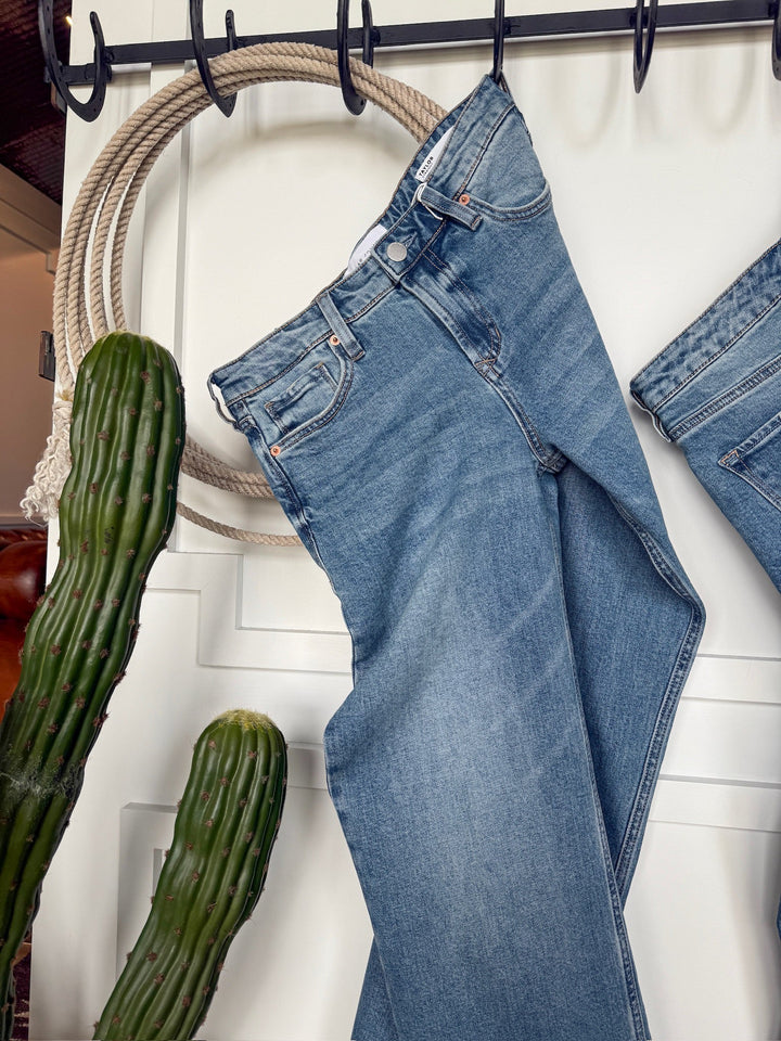 The Lennox Premier Rise Relaxed Straight Leg Jean by Savannah Sevens western life{&}style in light blue hangs by its belt loops on black hooks against a white wall, beside a coiled rope and a tall green cactus.
