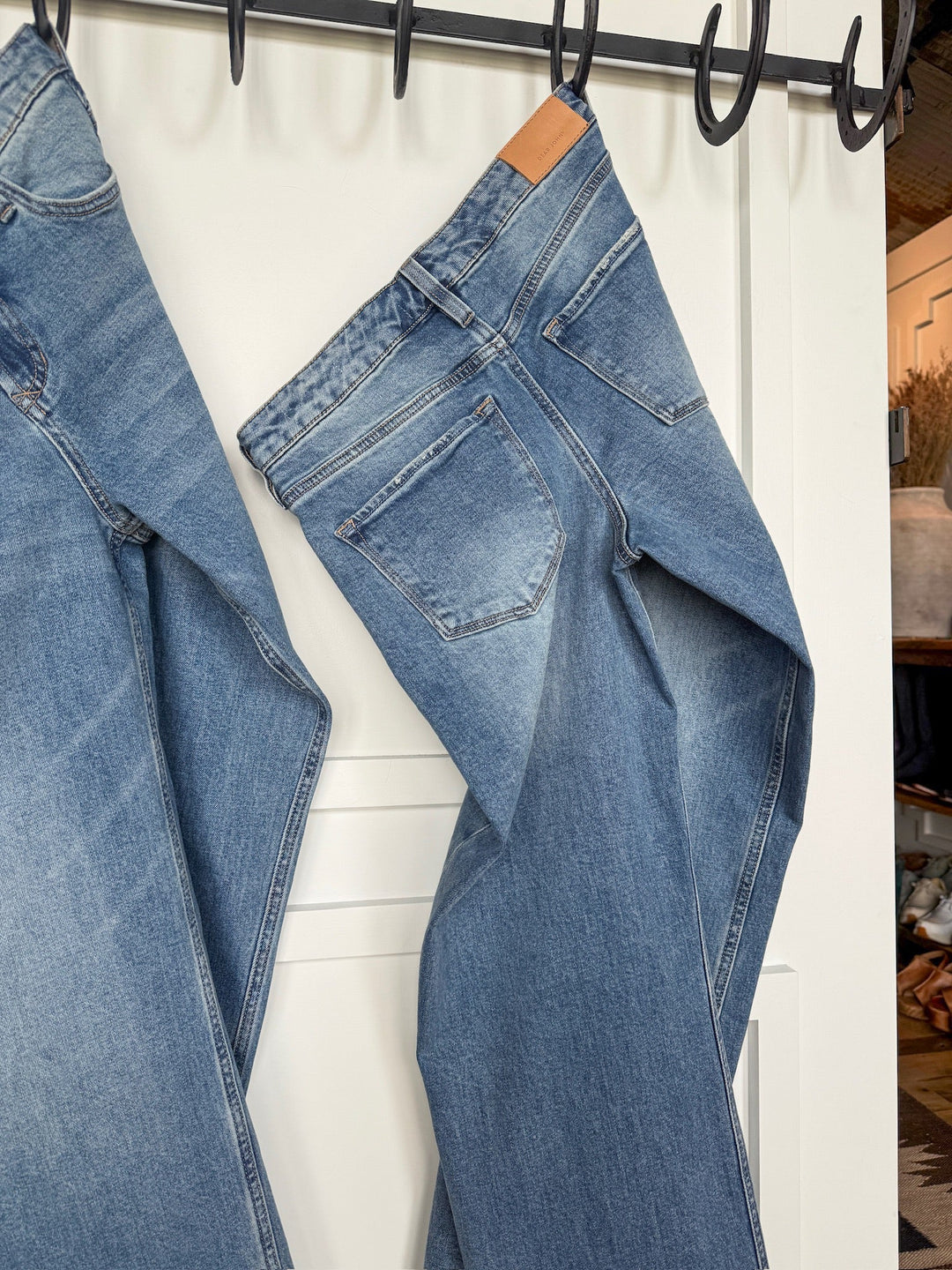 A pair of Savannah Sevens western life{&}style The Lennox Premier Rise Relaxed Straight Leg Jeans hangs by the waistband on a black hook rack attached to a white door, with another relaxed straight leg jean partially visible beside them.