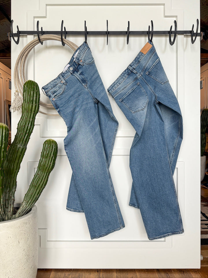 Two pairs of Savannah Sevens western life{&}style The Lennox Premier Rise Relaxed Straight Leg Jeans hang side by side on a black hook rack against a white wall, with a potted cactus and coiled rope in the background.
