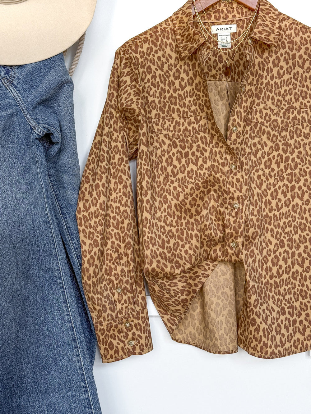 The Lenoir Leopard Button Up Top by Savannah Sevens western life{&}style, featuring long sleeves and a classic button-up front, is displayed on a hanger alongside blue jeans and a beige hat against a white background.
