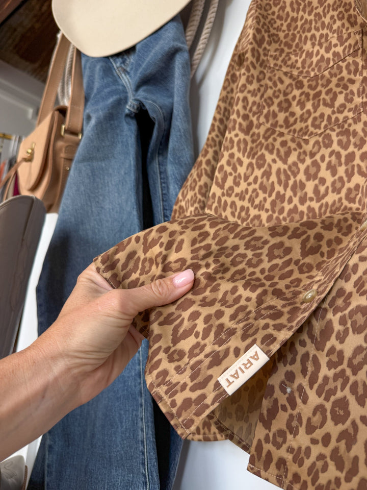A hand holds the sleeve of the Lenoir Leopard Button Up Top by Savannah Sevens western life{&}style. Beside it, blue jeans, a beige hat, a tan purse, and a taupe handbag are arranged on a white surface.