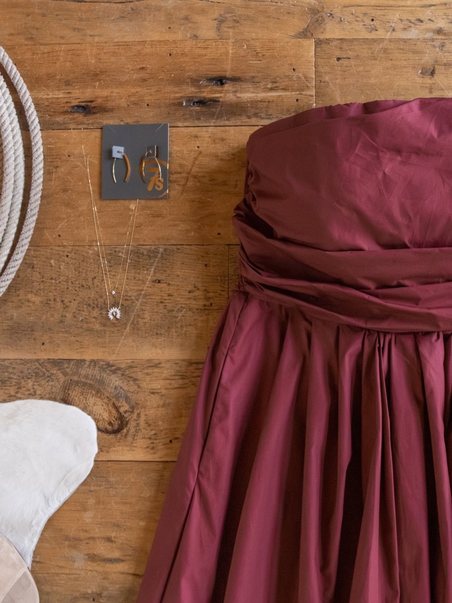 The Lenora Wine Strapless Dress by Savannah Sevens western life{&}style is displayed with two necklaces and two pairs of earrings on a wooden floor beside a white cowhide and a soft white hat.