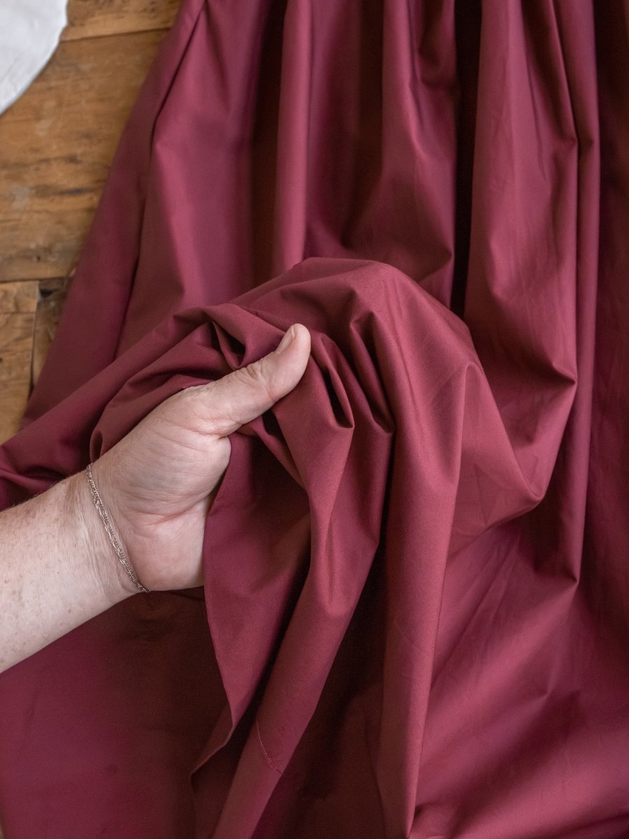 A hand gathers the rich fabric of the Lenora Wine Strapless Dress by Savannah Sevens western life{&}style, spread across a wooden surface.