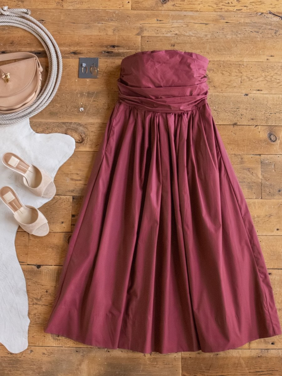 The Lenora Wine Strapless Dress by Savannah Sevens western life{&}style is displayed on a wooden floor with a beige handbag, white heeled shoes, and a coiled rope, partly resting on a white cowhide rug.