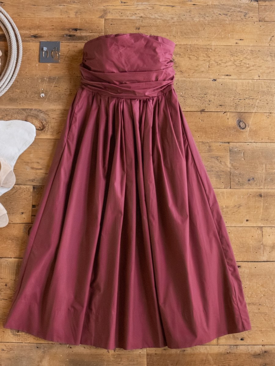 The Lenora Wine Strapless Dress by Savannah Sevens western life{&}style, featuring a fitted bodice and gathered skirt, is laid out on a wooden floor with silver earrings, a necklace, and a white accessory in the corner.