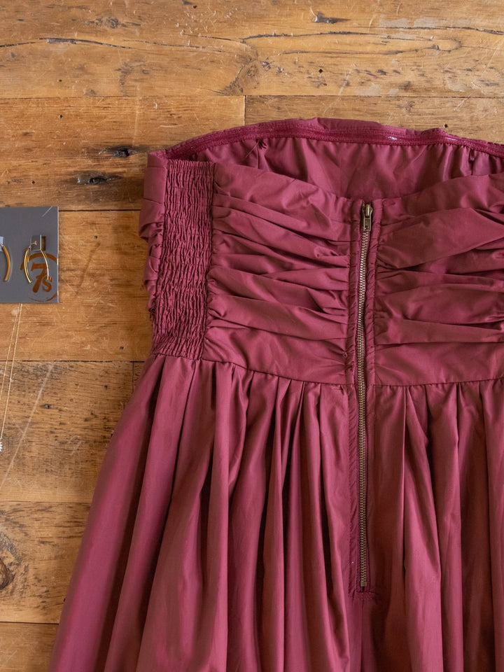 A close-up of the Lenora Wine Strapless Dress by Savannah Sevens western life{&}style with a ruched bodice, zipper closure, and smocked sides, laid flat beside a card featuring gold earrings.
