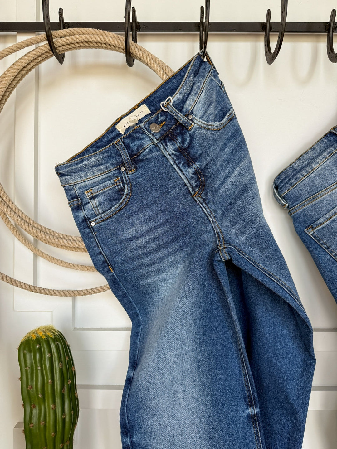 The Letty High Rise Wide Leg Jean by Savannah Sevens western life{&}style hangs on a black hook against a white wall, with a coiled rope and cactus nearby. Another pair of wide leg jeans is partially visible to the right.
