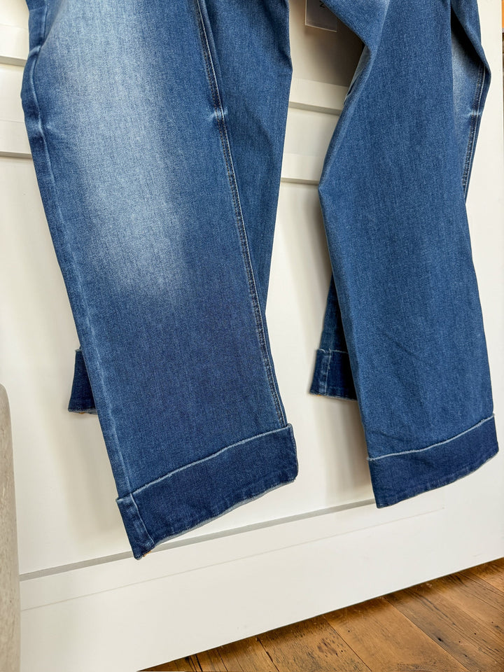 A close-up of two pairs of The Letty High Rise Wide Leg Jean by Savannah Sevens western life{&}style, medium wash denim with folded cuffs, hanging side by side against a white background and wooden floor.