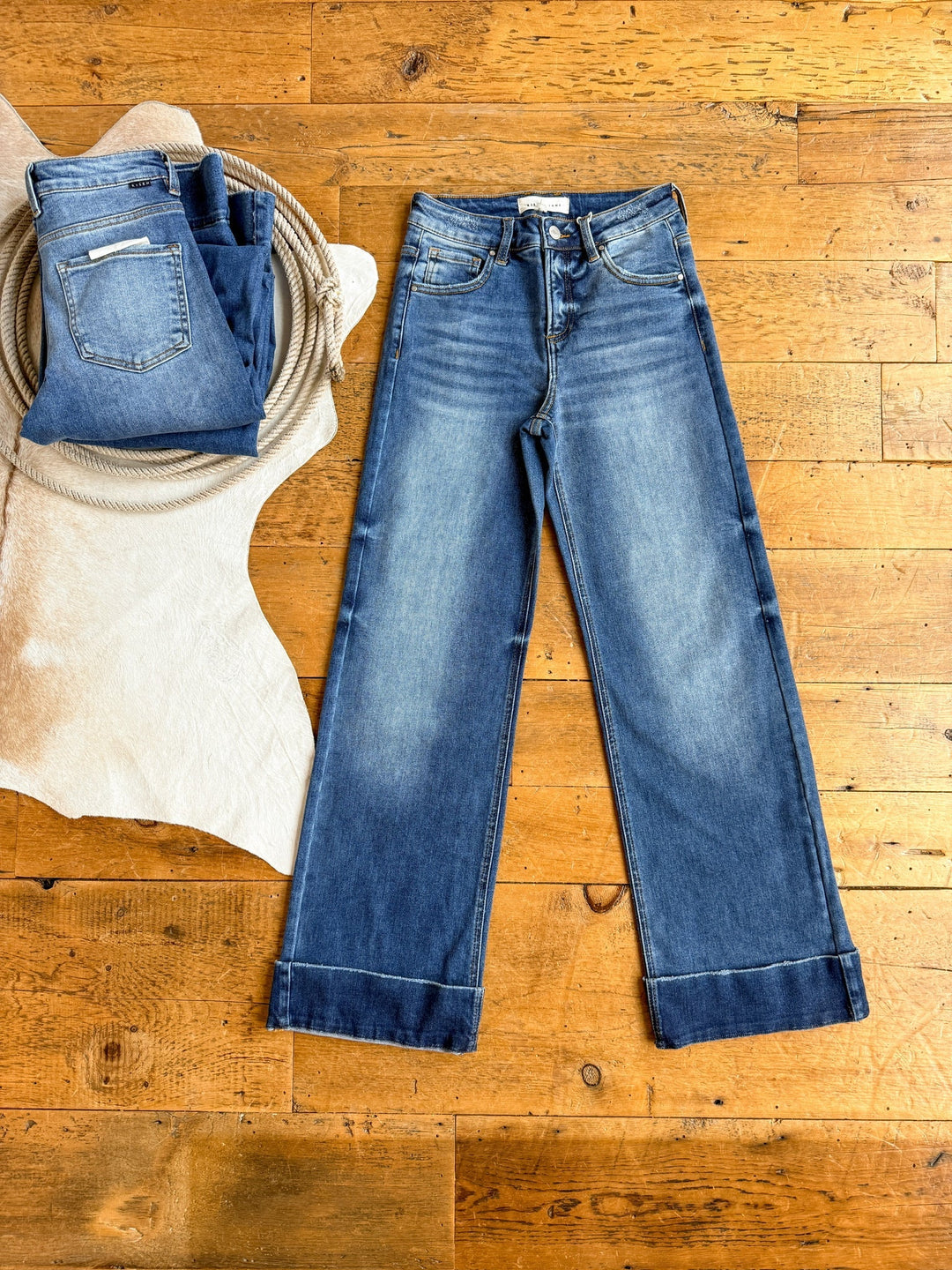 The Letty High Rise Wide Leg Jean by Savannah Sevens western life{&}style is laid flat on a wooden floor, beside a folded medium wash denim Risen Jeans on white textured fabric with coiled rope.