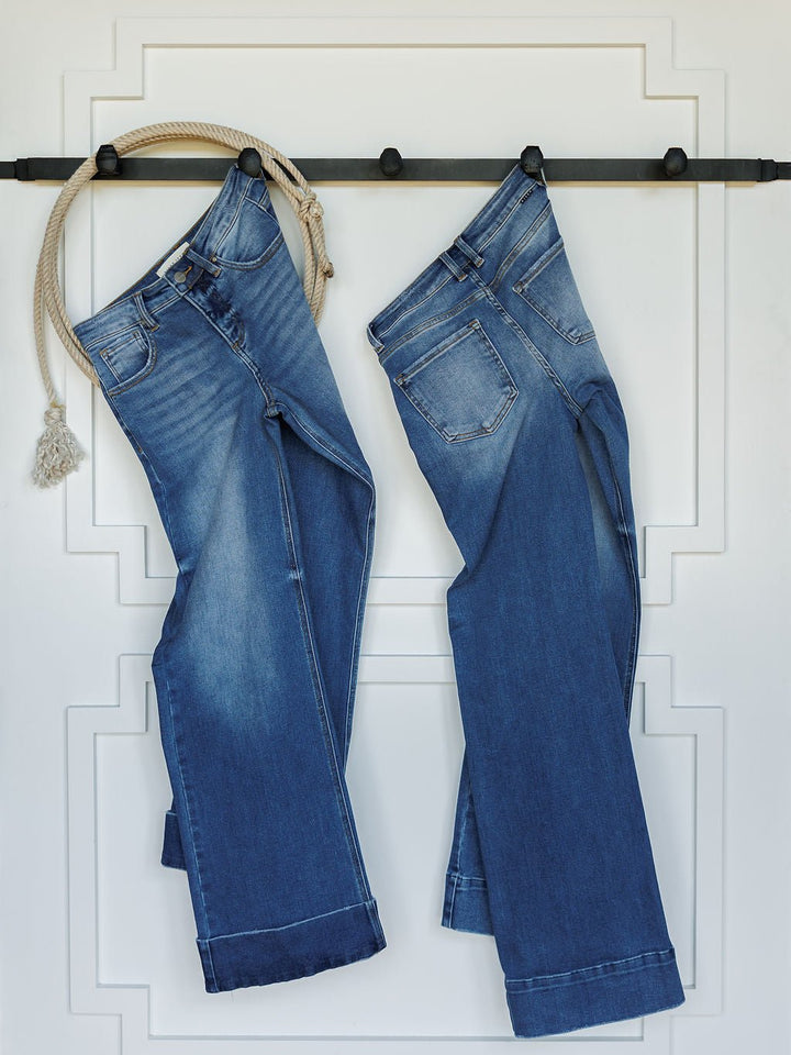 Two pairs of The Letty High Rise Wide Leg Jeans by Savannah Sevens western life{&}style hang side by side on black hooks against a white paneled wall, with a beige rope tassel looped through the left pair’s belt loop.