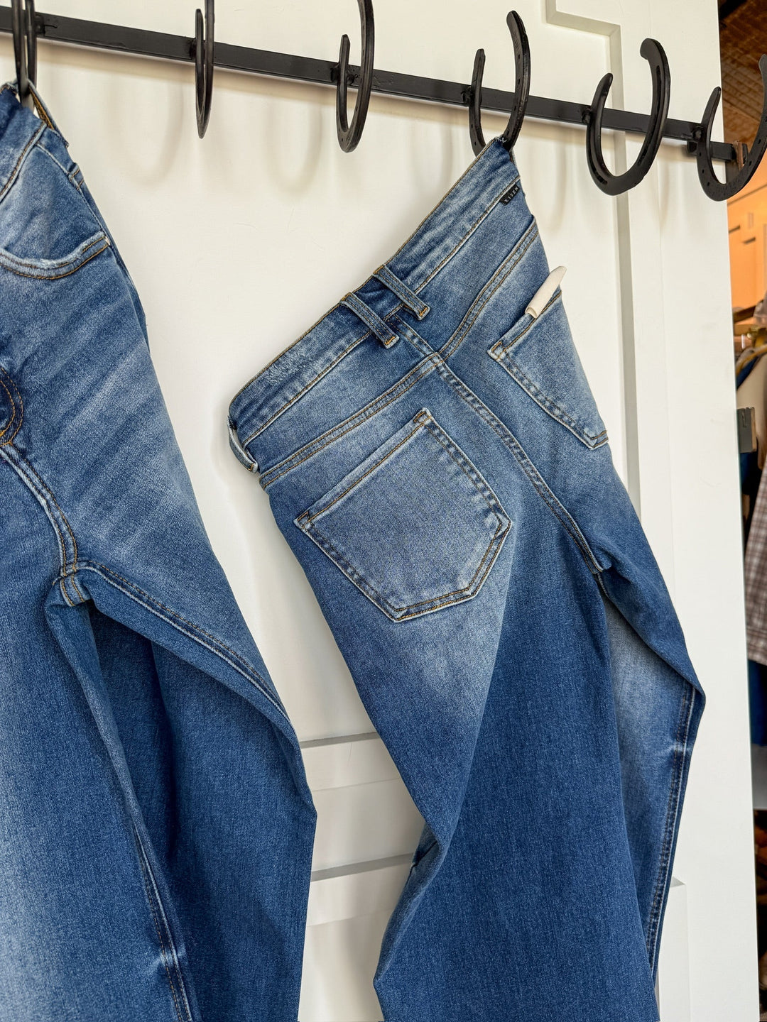 A pair of Savannah Sevens western life{&}style The Letty High Rise Wide Leg Jeans in medium wash denim hang by their belt loops on a black metal hook rack attached to a white wall, with another pair of Risen Jeans partially visible next to them.