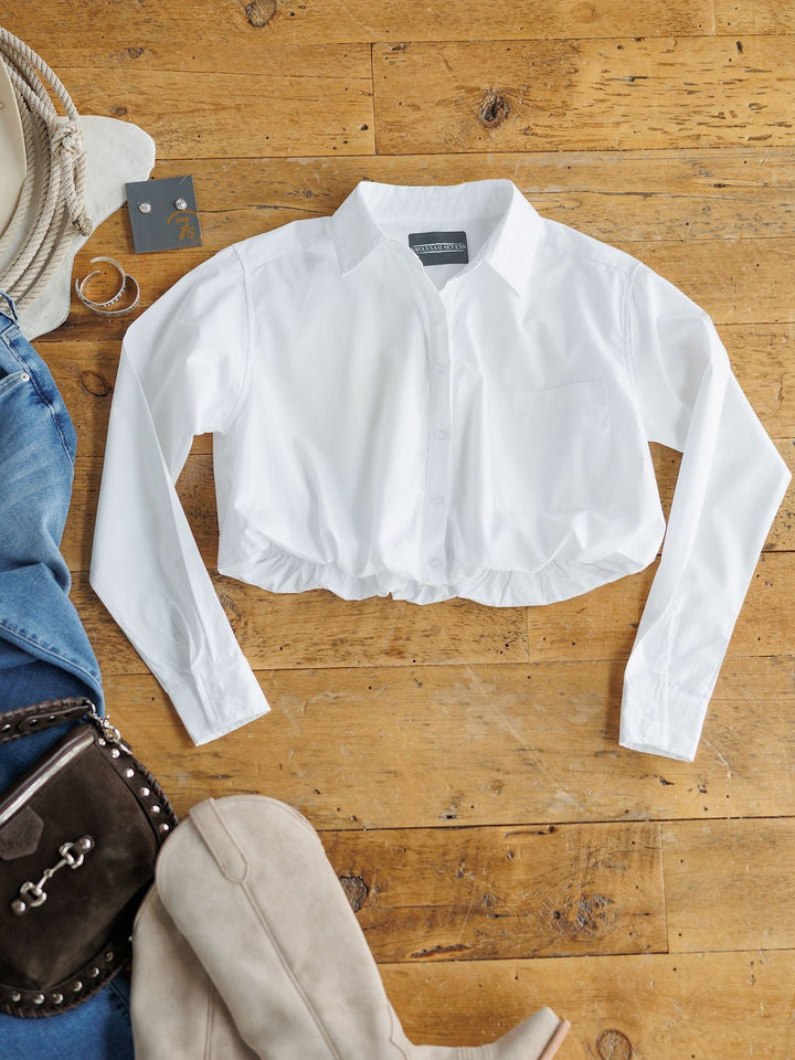 The Levasy White Bubble Hem Cropped Button Up Top by Savannah Sevens western life{&}style is styled flat with jeans, a  bag, boots, silver hoop earrings, and sunglasses on a wooden floor.