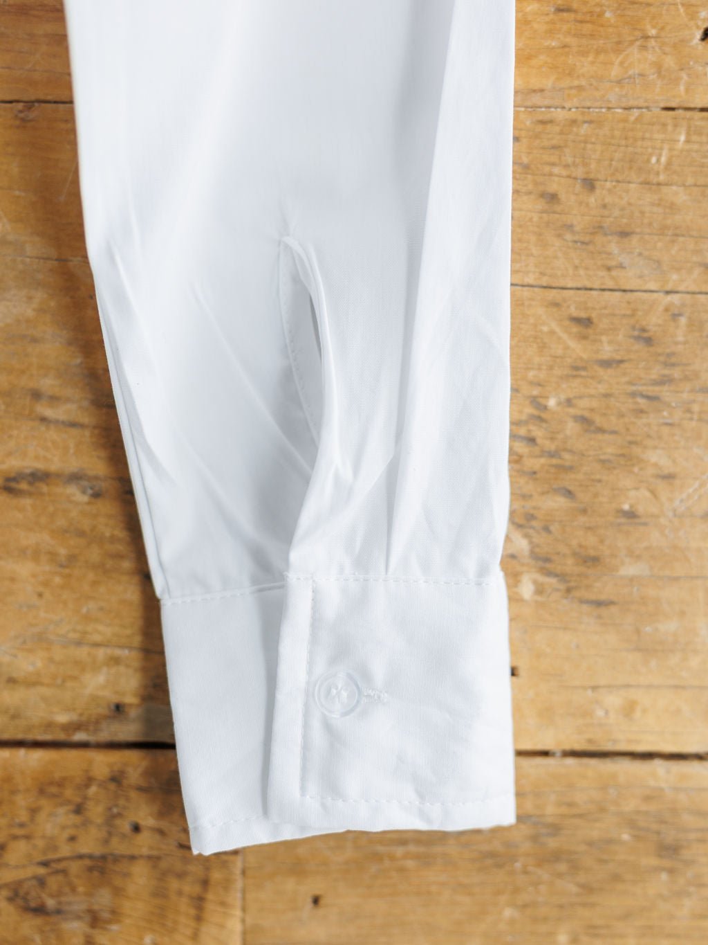 A close-up of the Levasy White Bubble Hem Cropped Button Up Top by Savannah Sevens western life{&}style shows its buttoned cuff and smooth, neatly pressed cotton fabric resting on a wooden surface.
