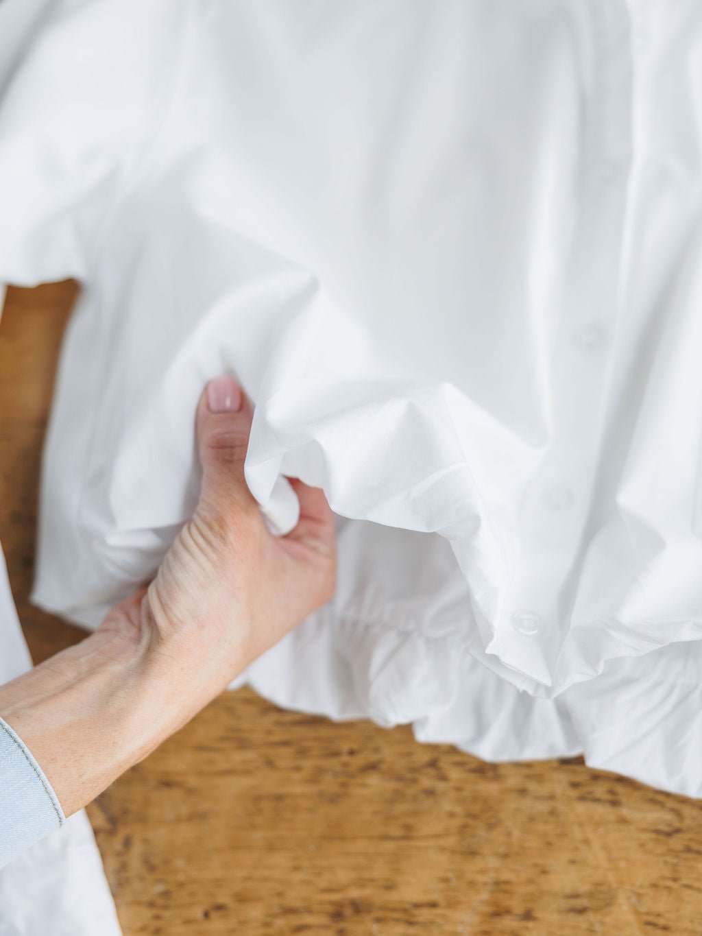 A hand delicately holds the Levasy White Bubble Hem Cropped Button Up Top by Savannah Sevens western life{&}style, showcasing its ruffled hem over a wooden surface.