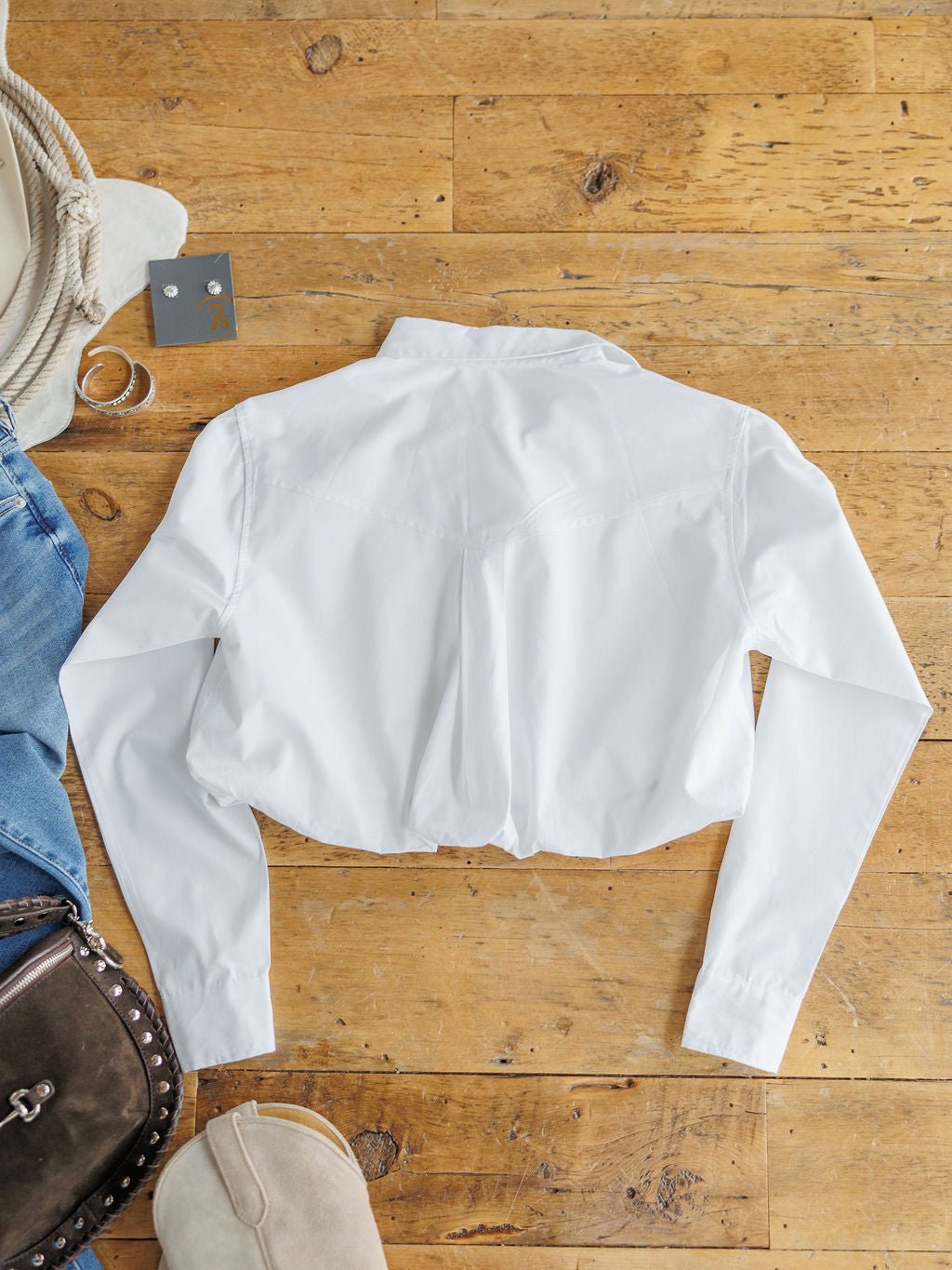 The Levasy White Bubble Hem Cropped Button Up Top by Savannah Sevens western life{&}style is laid flat on a wooden floor, surrounded by jeans, a brown leather bag, beige shoes, a coiled rope, and silver ring.