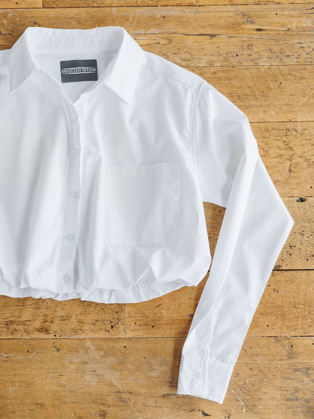 The Levasy White Bubble Hem Cropped Button Up Top by Savannah Sevens western life{&}style, featuring long sleeves, a collar, and chest pocket, is displayed on a wooden surface.