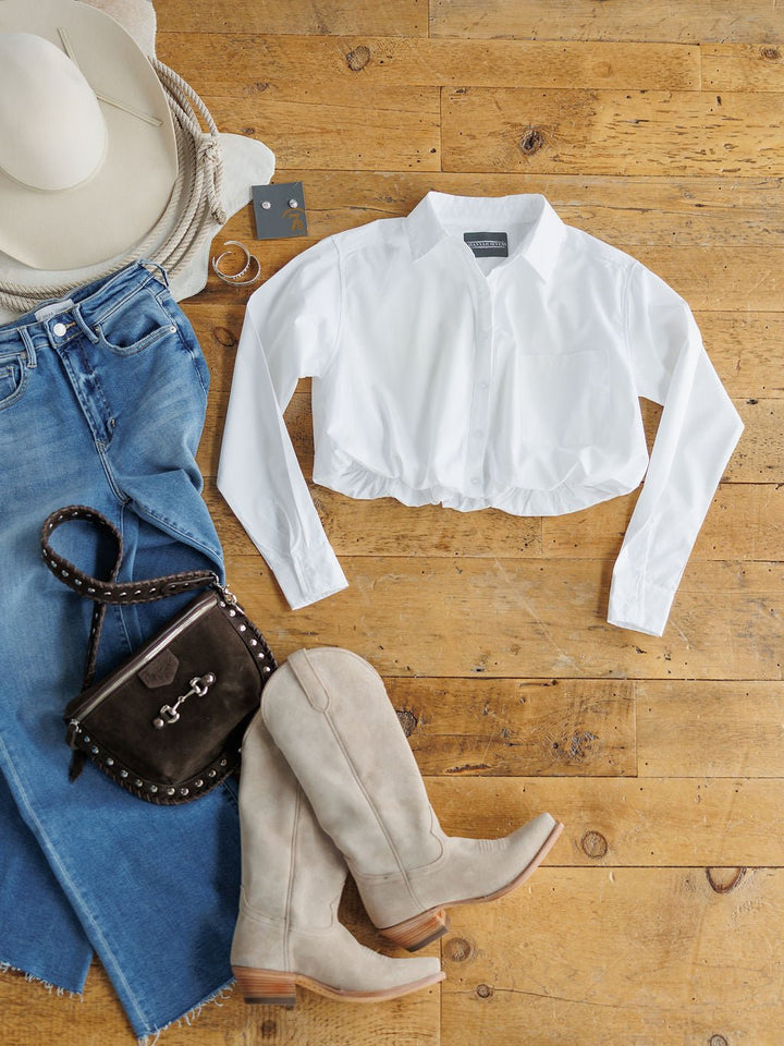 Flat lay of the Levasy White Bubble Hem Cropped Button Up Top by Savannah Sevens western life{&}style, blue jeans, beige knee-high boots, black studded purse, white hat, hoop earrings, and a silver ring on a wooden floor.