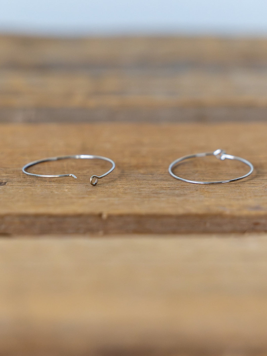 Two thin Levy Gold/Silver Hoop Earrings by Savannah Sevens western life{&}style rest on a wooden surface, with one hoop open and the other closed. Both are tarnish resistant.