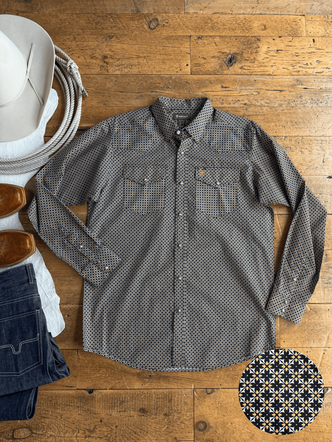 A Lewisburg Ariat Men’s Southwest Pearl Snap shirt from Savannah Sevens western life{&}style lies on a wooden floor, surrounded by brown boots, a hat, a rope, and denim jeans. An inset displays a close-up of the shirt’s black, white, and tan southwest pattern print.