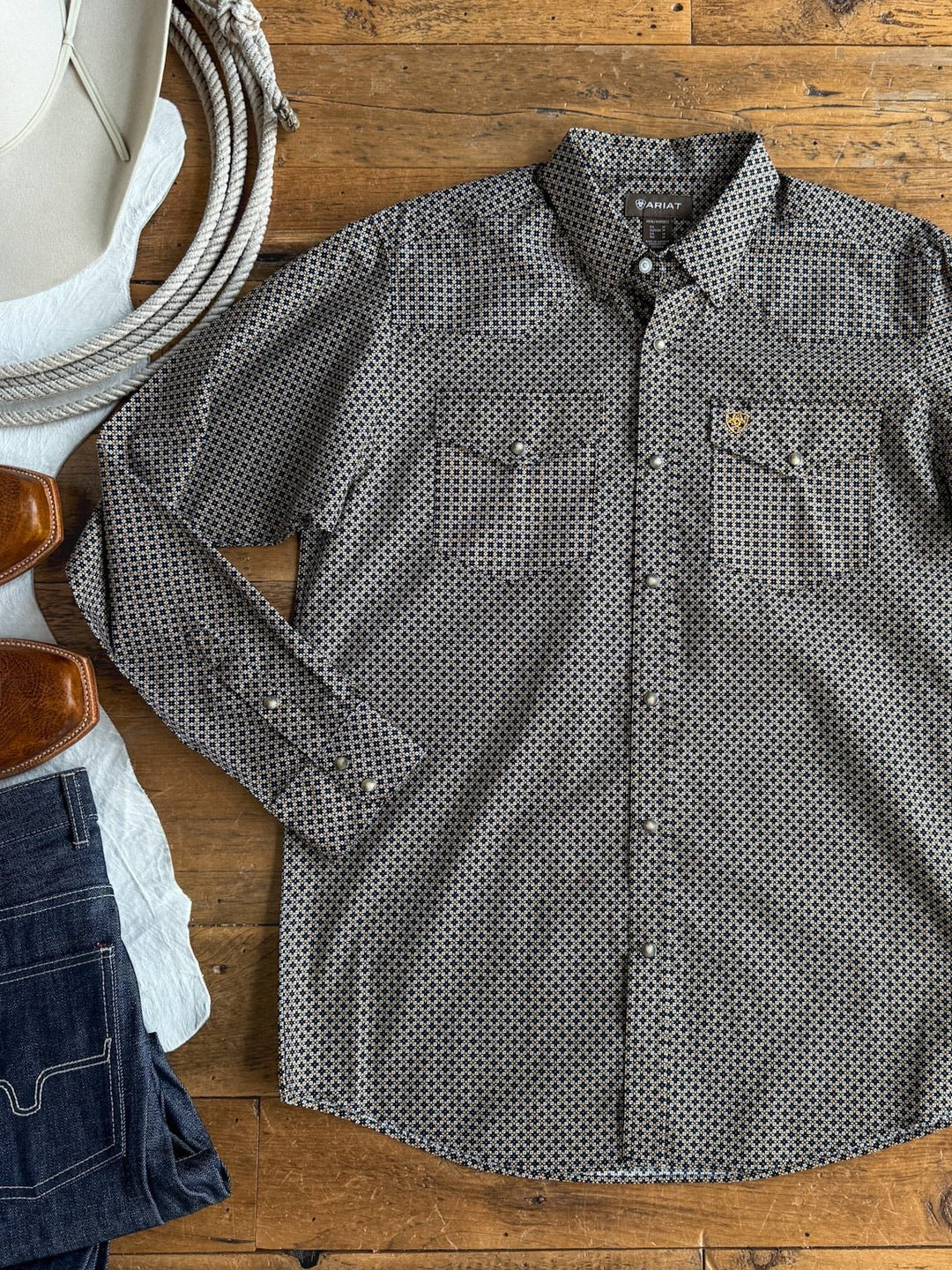 A Lewisburg Ariat Men’s Southwest Pearl Snap shirt from Savannah Sevens western life{&}style is laid flat on a wooden floor next to a cowboy hat, brown boots, and folded denim jeans.