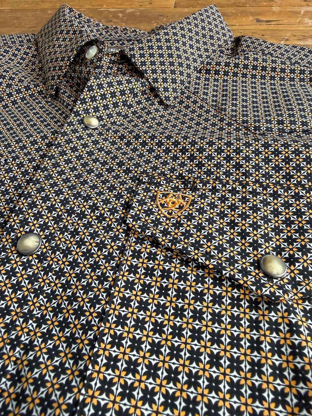 Close-up of the Lewisburg Ariat Men’s Southwest Pearl Snap by Savannah Sevens western life{&}style—a long sleeve shirt featuring black, white, and yellow southwest-inspired geometric designs. The shirt is adorned with silver pearl snap buttons and a small orange embroidered logo on the chest pocket, and is displayed laid out on a wooden surface.