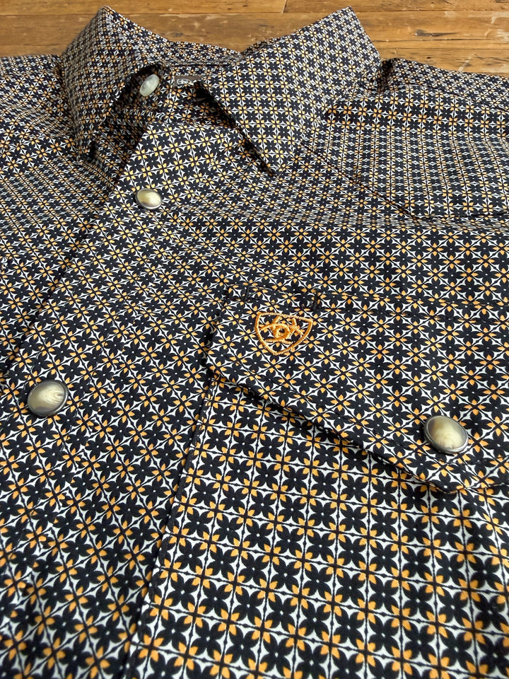 Close-up of the Lewisburg Ariat Men’s Southwest Pearl Snap by Savannah Sevens western life{&}style—a long sleeve shirt featuring black, white, and yellow southwest-inspired geometric designs. The shirt is adorned with silver pearl snap buttons and a small orange embroidered logo on the chest pocket, and is displayed laid out on a wooden surface.