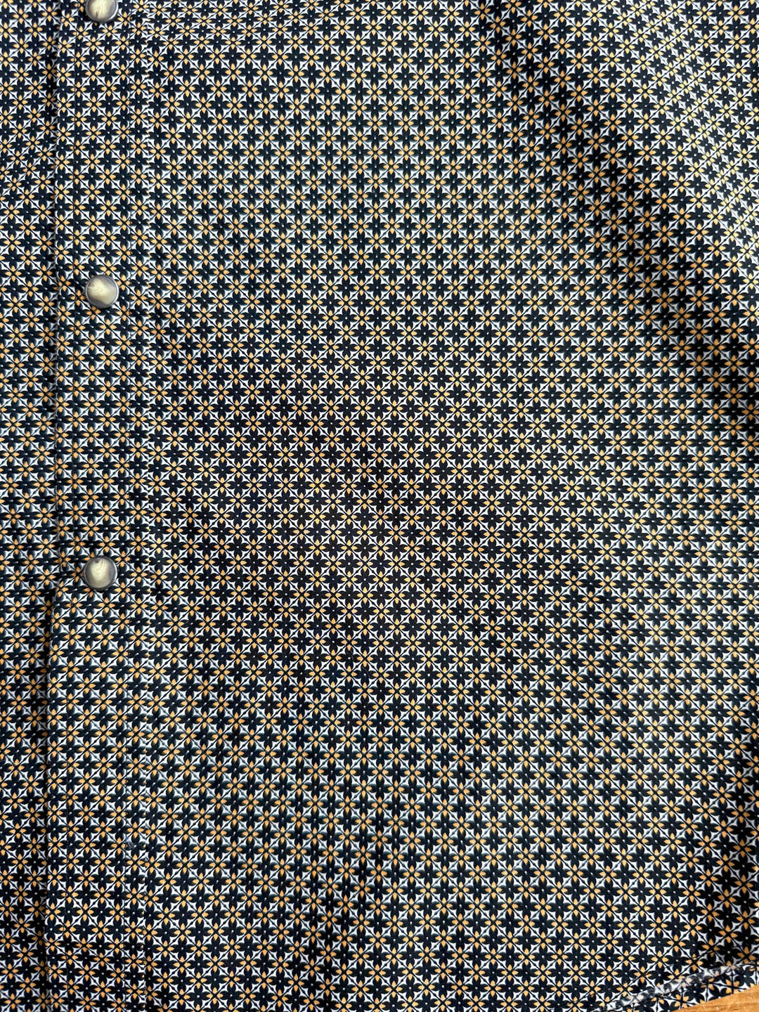 A close-up of the Lewisburg Ariat Men’s Southwest Pearl Snap shirt by Savannah Sevens western life{&}style, showcasing its small geometric blue, white, and yellow pattern and silver snap buttons along the left edge.