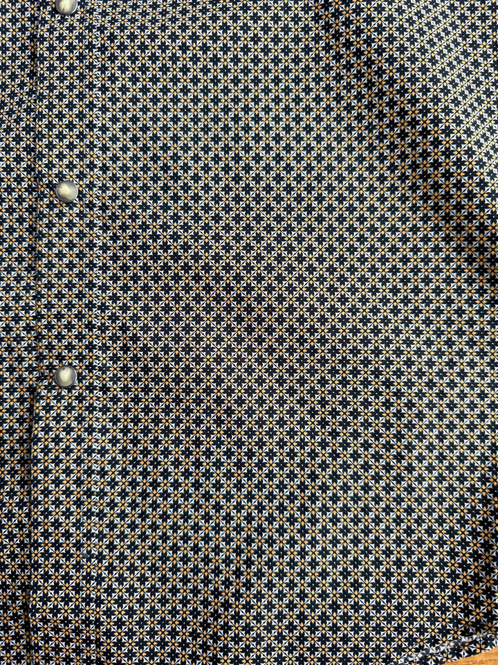 A close-up of the Lewisburg Ariat Men’s Southwest Pearl Snap shirt by Savannah Sevens western life{&}style, showcasing its small geometric blue, white, and yellow pattern and silver snap buttons along the left edge.