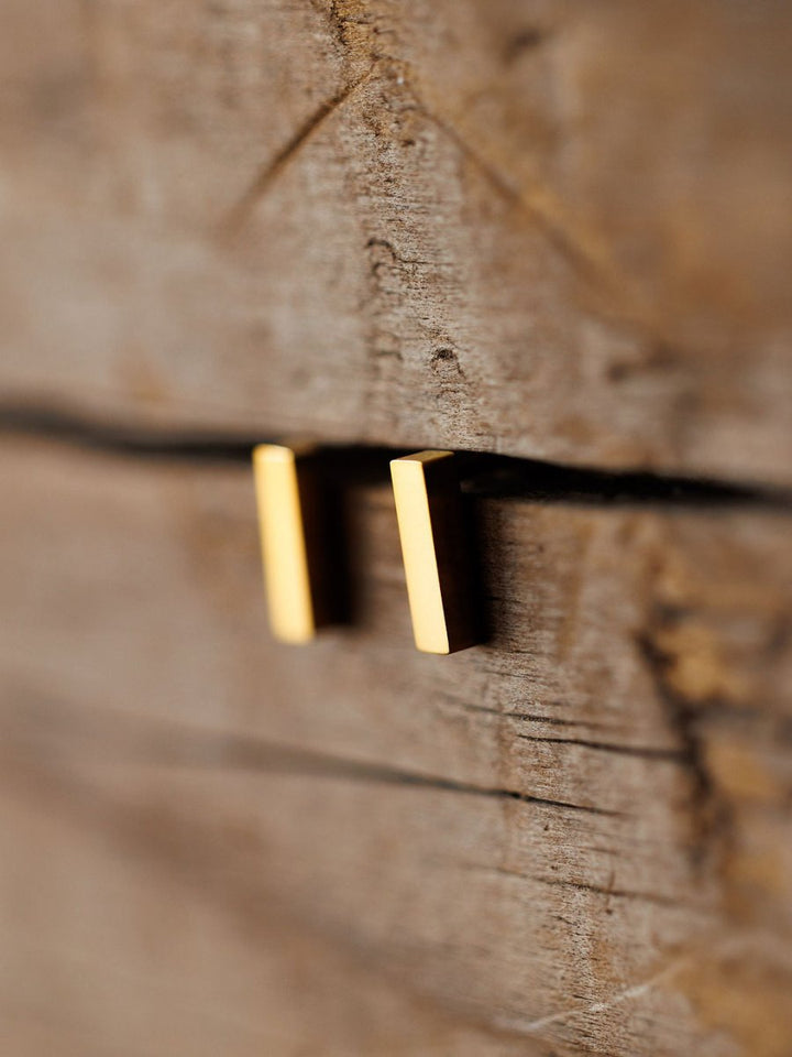 A close-up of the Lexington Earrings by Savannah Sevens western life{&}style—minimalist gold-coated bar studs—displayed vertically on a textured wooden background.