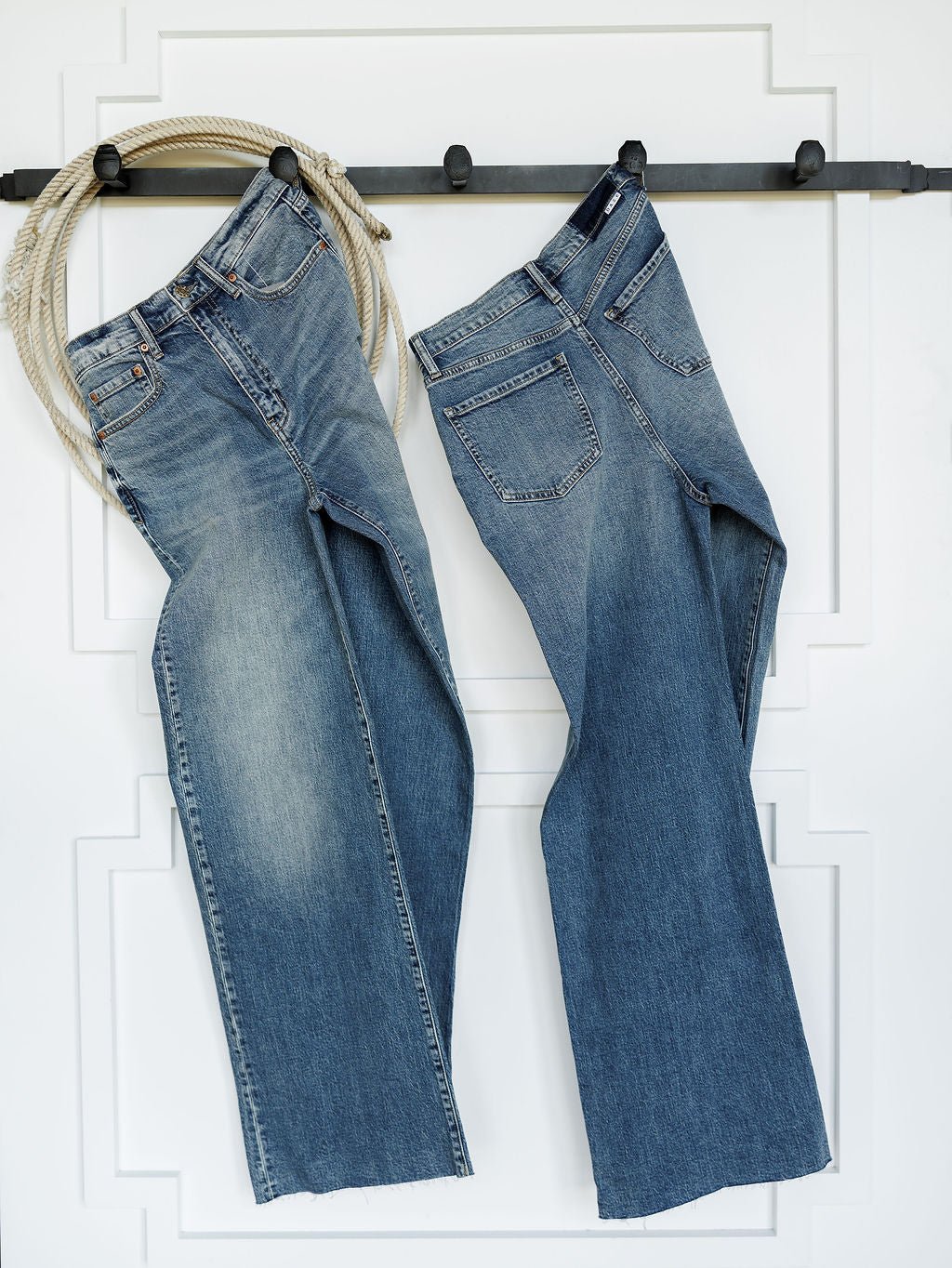 Two pairs of The Leyden High Rise Wide Leg Jean by Savannah Sevens western life{&}style hang on a black wall hook, with a coiled rope draped over the left hook. A white wall with decorative molding serves as the background.